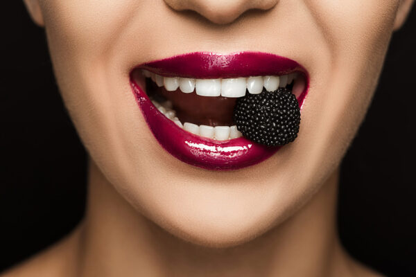 Nahaufnahme eines lächelnden Mundes mit glänzendem, pinkfarbenem Lippenstift, der eine schwarze Süßigkeit zwischen den Zähnen hält.